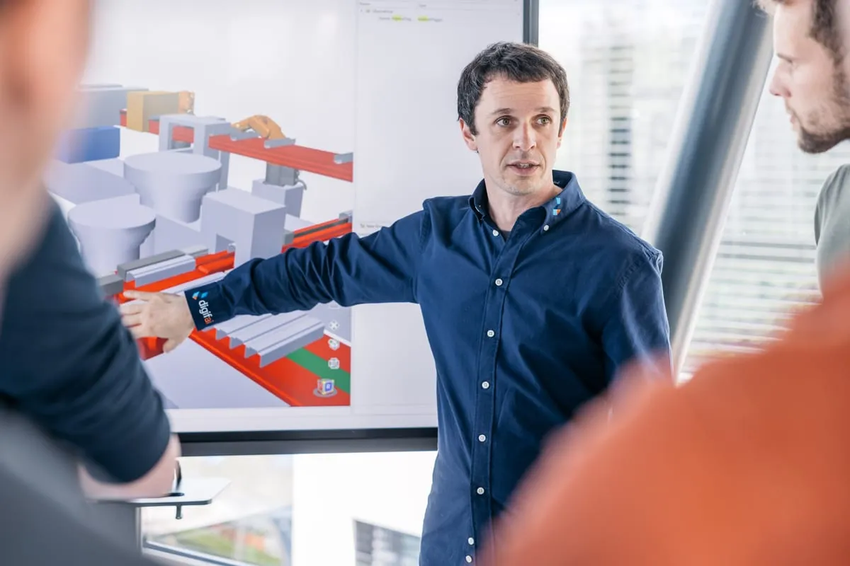 A digifai engineer explains the digital twin of a production line with robot and conveyor to colleagues on a large screen.